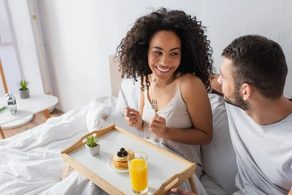 Happy african american woman holding cutlery and looking at boyfriend in bedroom — Stock Photo