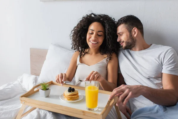 Happy african american woman holding cutlery near pancakes and boyfriend in bedroom — Stock Photo