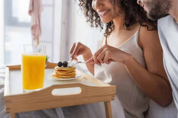 Cropped view of happy african american woman holding cutlery near pancakes and boyfriend in bedroom — Stock Photo