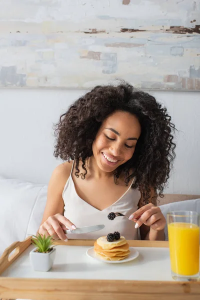 Mujer afroamericana feliz sosteniendo tenedor con mora cerca de panqueques en bandeja - foto de stock