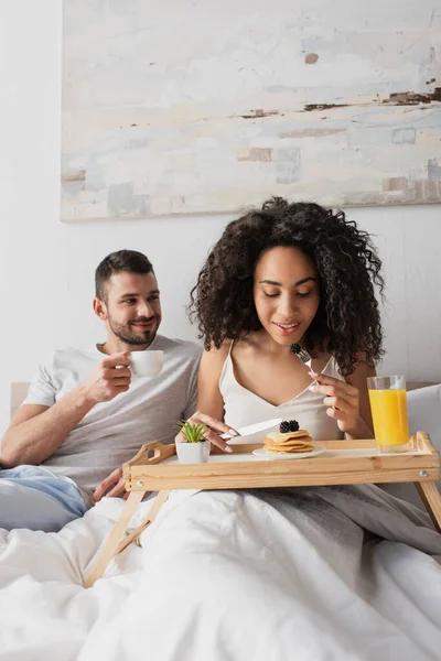 Happy african american woman holding fork with blackberry near pancakes on tray and boyfriend with cup of coffee — Stock Photo