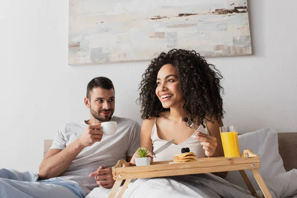 Femme afro-américaine souriante tenant fourchette avec mûre près de crêpes sur plateau et petit ami avec tasse de café — Photo de stock