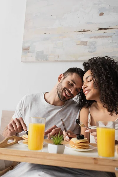 Cheerful interracial couple smiling near breakfast on tray in bedroom — Stock Photo