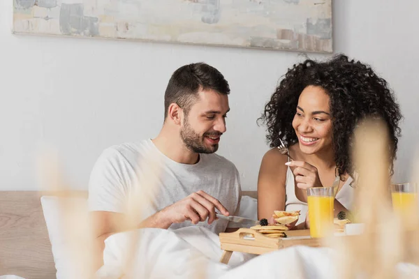 Alegre pareja interracial sonriendo y desayunando en el dormitorio - foto de stock