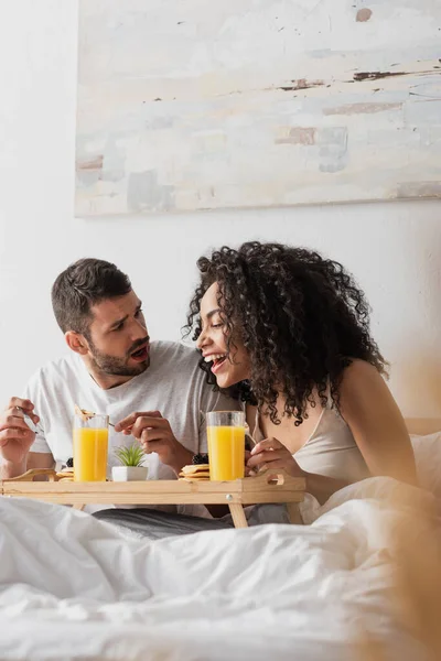 Шокированный бородатый мужчина смотрит на счастливую африканскую американскую подружку, завтракающую — стоковое фото