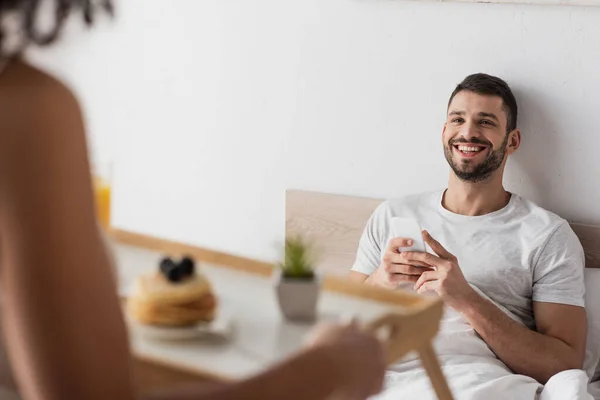 Hombre feliz con teléfono inteligente mirando a la novia afroamericana sosteniendo bandeja con desayuno en primer plano borrosa - foto de stock
