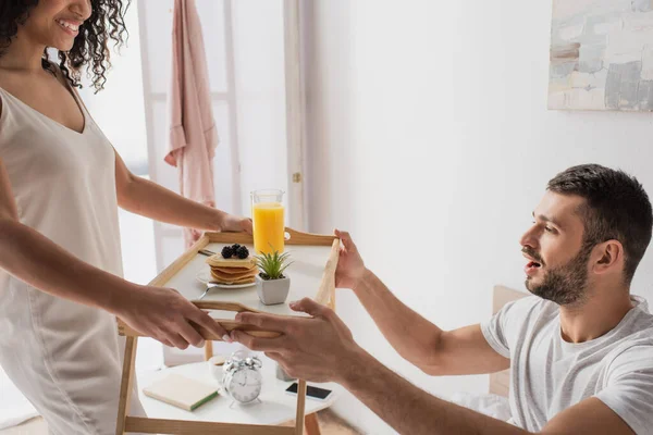Happy african american woman giving tray with breakfast to cheerful boyfriend — Stock Photo