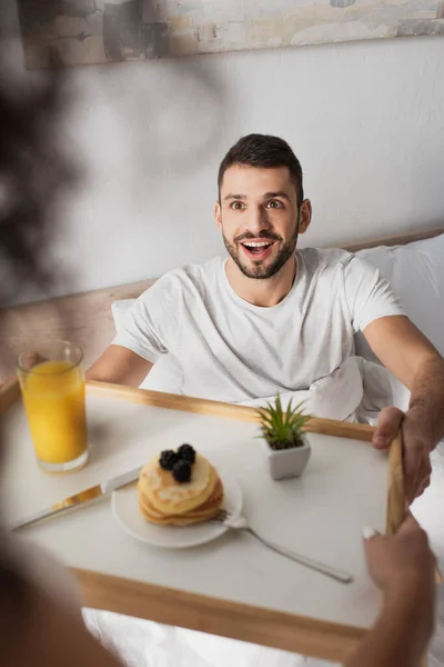 Amazed man taking tray with breakfast from hands of african american girlfriend — Stock Photo
