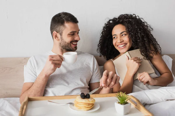 Fröhliche afrikanisch-amerikanische halten Buch in der Nähe glücklichen Freund beim Frühstück im Bett — Stockfoto