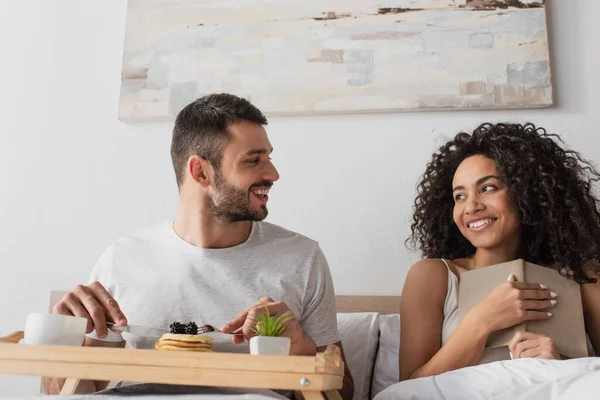 Happy african american holding book and looking at boyfriend having breakfast in bed — Stock Photo