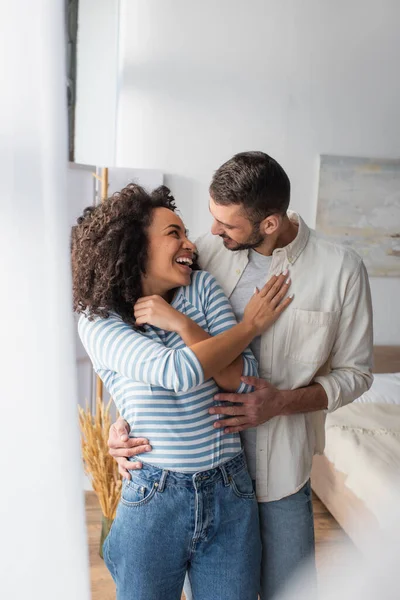 Радісна багатоетнічна пара сміється, обіймаючись вдома — стокове фото