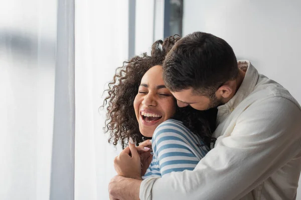 Alegre casal multiétnico abraçando em casa — Fotografia de Stock