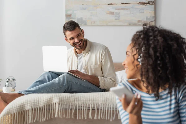 Glücklicher Mann mit Laptop in der Nähe einer afrikanisch-amerikanischen Frau in drahtlosen Kopfhörern, die Musik auf verschwommenem Hintergrund hört — Stockfoto