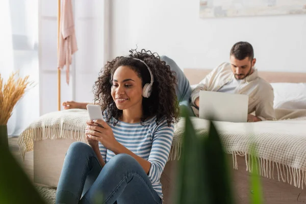 Cheerful african american woman in wireless headphones holding smartphone near boyfriend using laptop on blurred background — Stock Photo