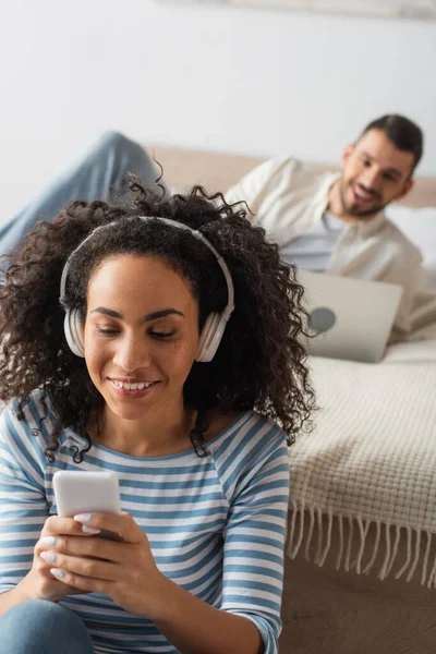 Sorridente mulher afro-americana em fones de ouvido sem fio segurando smartphone perto do namorado usando laptop em fundo borrado — Fotografia de Stock
