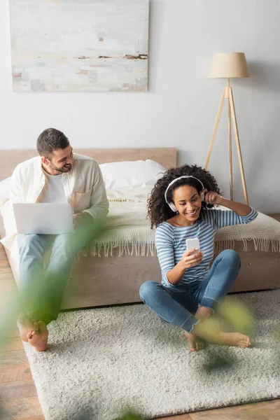 Sonriente mujer afroamericana en auriculares inalámbricos sosteniendo teléfono inteligente cerca de novio usando portátil con primer plano borroso - foto de stock