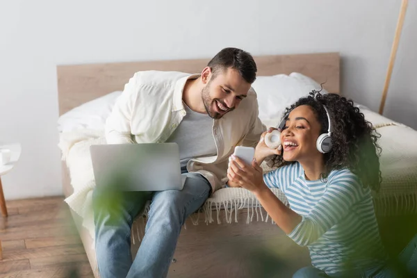 Happy african american woman in wireless headphones holding smartphone near smiling boyfriend with laptop — Stock Photo