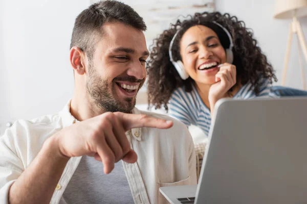 Hombre feliz señalando con el dedo a la computadora portátil cerca de la mujer afroamericana en auriculares inalámbricos sobre fondo borroso - foto de stock