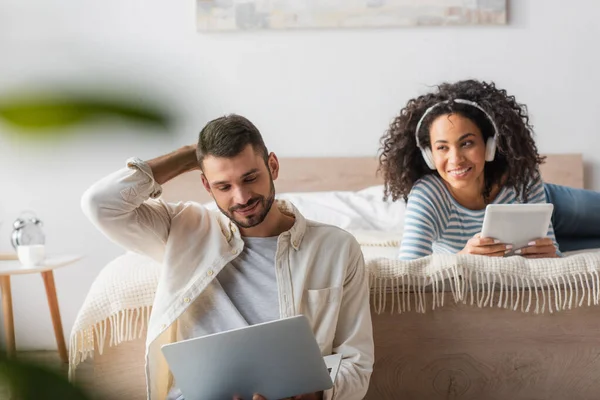 Bärtiger Mann mit Laptop in der Nähe einer Afroamerikanerin mit drahtlosen Kopfhörern auf verschwommenem Hintergrund — Stockfoto