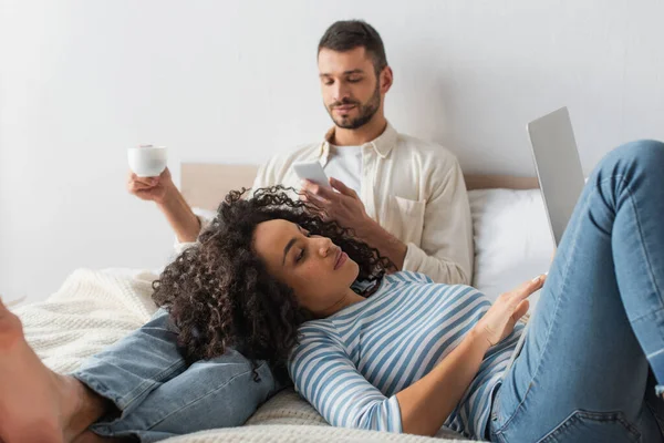 Mulher afro-americana deitada na cama e usando laptop perto namorado com copo e smartphone — Fotografia de Stock