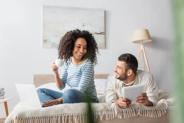 Heureuse femme afro-américaine assise sur le lit, tenant la tasse et utilisant un ordinateur portable près du petit ami avec tablette numérique — Photo de stock