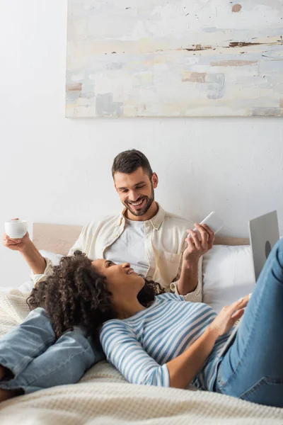 Sonriente afroamericana mujer acostada en la cama y el uso de la computadora portátil cerca de novio con taza y teléfono inteligente - foto de stock