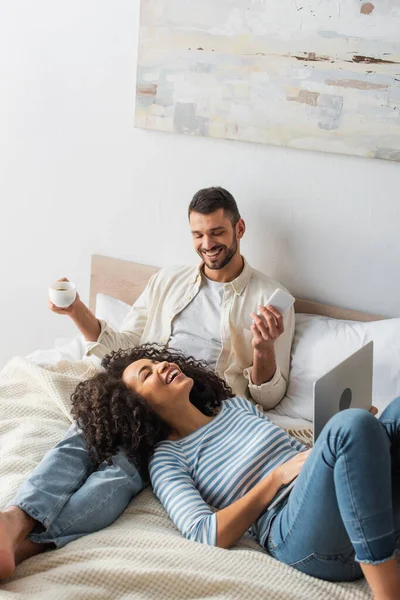 Mulher americana africana feliz deitado na cama e usando laptop perto namorado com copo e smartphone — Fotografia de Stock
