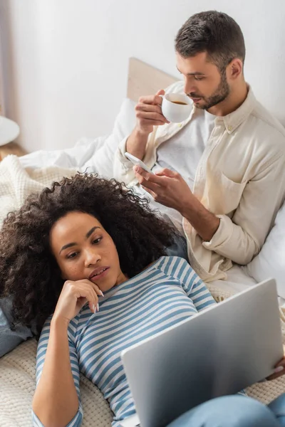 Vista ad alto angolo della donna afro-americana sdraiata sul letto e utilizzando laptop vicino al fidanzato con smartphone bere caffè — Foto stock