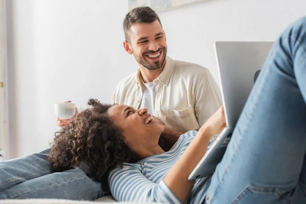 Happy african american woman lying on bed and laughing near laptop and boyfriend — Stock Photo