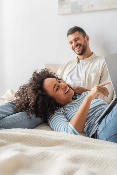 Happy african american woman lying on bed and pointing with finger at laptop near boyfriend — Stock Photo