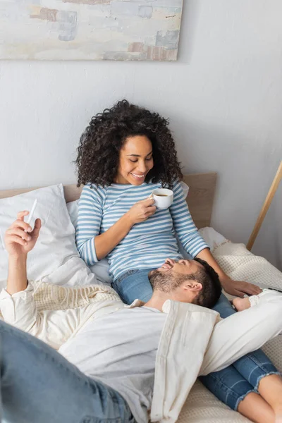 Glückliche Afroamerikanerin hält Tasse neben bärtigen Freund, der mit Smartphone auf Bett liegt — Stockfoto