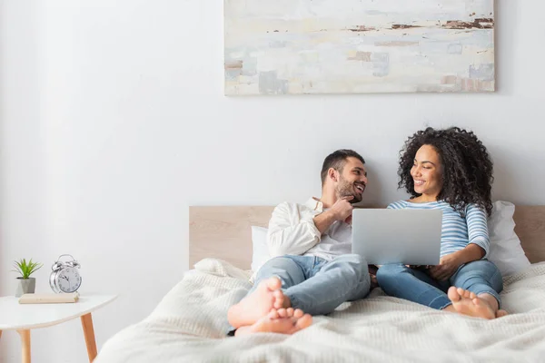 Feliz casal interracial refrigeração na cama e assistir filme no laptop — Fotografia de Stock