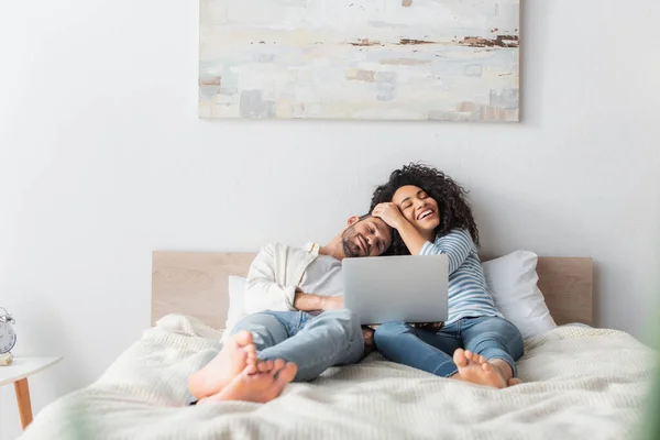 Межрасовая пара улыбается и отдыхает в постели во время просмотра фильма на ноутбуке — стоковое фото