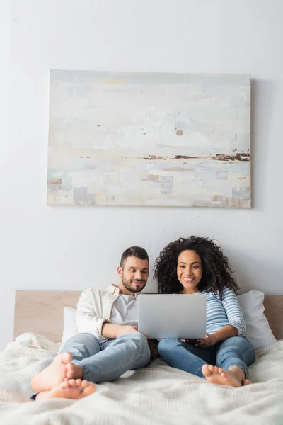 Feliz interracial pareja chilling en cama y viendo comedia película en laptop - foto de stock