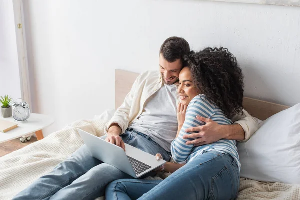 Alegre interracial pareja chilling en cama y viendo comedia película en laptop en dormitorio - foto de stock