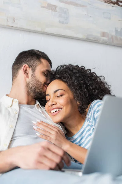 Hombre besando feliz afroamericano novia mientras se enfría en la cama con el ordenador portátil - foto de stock