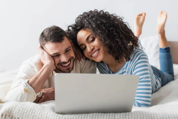 Alegre interracial casal refrigeração na cama enquanto assiste filme no laptop — Fotografia de Stock