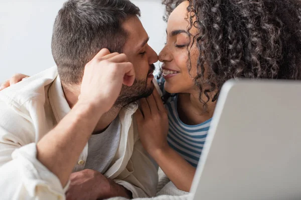 Pareja interracial besándose mientras se enfría en la cama cerca del portátil - foto de stock