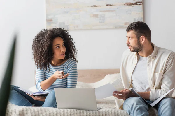 Mujer afroamericana mirando novio sosteniendo papeles con impuestos cerca de portátil en la cama - foto de stock