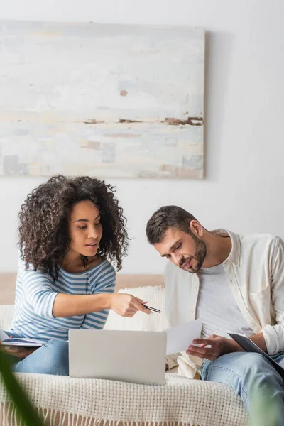 African american woman pointing at paper with taxes near boyfriend and laptop on bed — Stock Photo