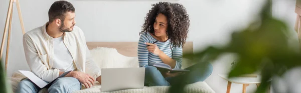 African american woman pointing at boyfriend sitting with folder near laptop on bed, banner — Stock Photo