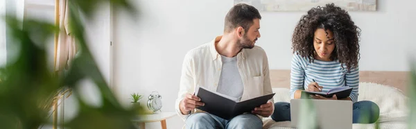 Barbudo hombre sosteniendo carpeta con impuestos y mirando a la novia afroamericana con cuaderno sentado en la cama, bandera — Stock Photo