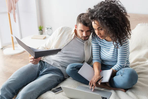 Mujer afroamericana con portátil usando portátil cerca novio barbudo celebración carpeta en el dormitorio - foto de stock