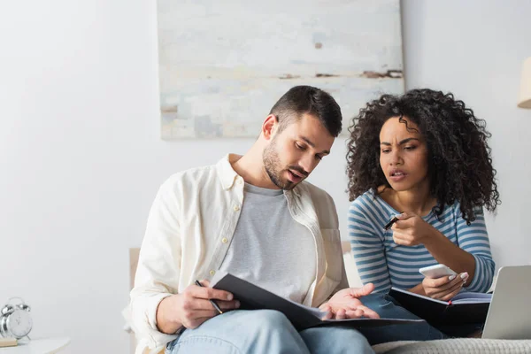 African american woman holding smartphone and pointing with pen at folder in hands of boyfriend — Stock Photo