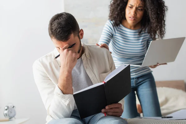 Hombre sosteniendo el cuaderno y cubriendo la cara cerca preocupada novia afroamericana sobre fondo borroso - foto de stock