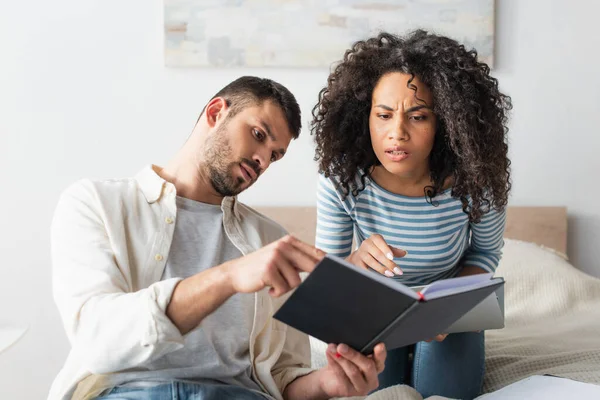 Pareja interracial mirando cuaderno en el dormitorio - foto de stock