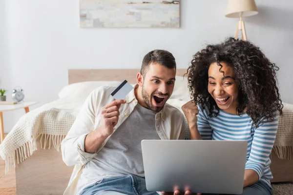 Amazed interracial couple looking at laptop while shopping online — Stock Photo