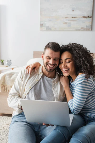 Feliz pareja interracial sonriendo mientras mira el ordenador portátil - foto de stock