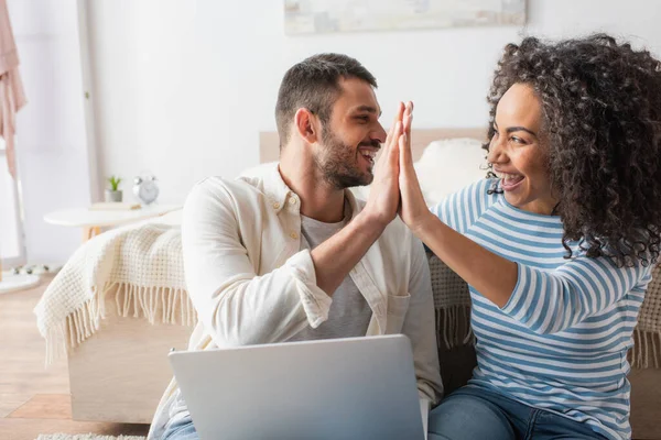 Heureux interracial couple sourire tout en donnant haute cinq près ordinateur portable — Photo de stock