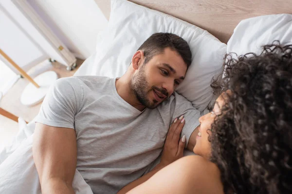 Vue grand angle de couple interracial positif se regardant tout en étant couché dans le lit à la maison — Photo de stock